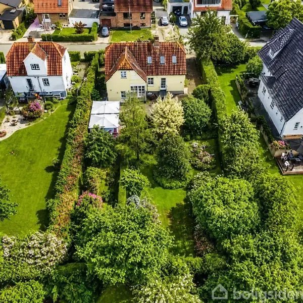 Huse ligger tæt på hinanden med velplejede haver fyldt med grønne træer og buske. De har røde og sorte tegltage, omgivet af rækkehuse og grønne områder i et villakvarter.