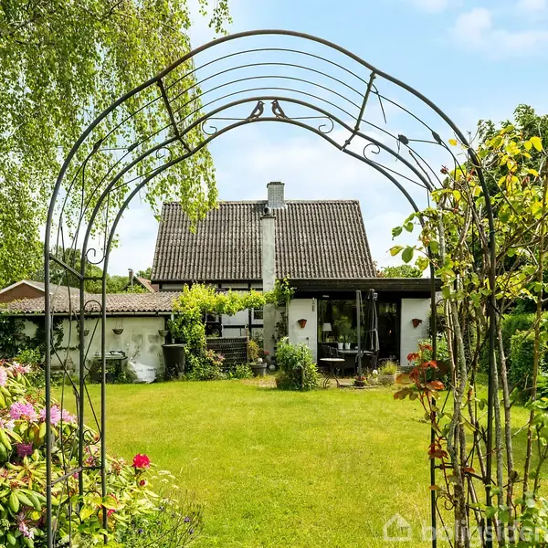 En metalbue står i en frodig have, dekoreret med klatrende planter; i baggrunden ses et hus med gråt tag omgivet af buske og blomster.