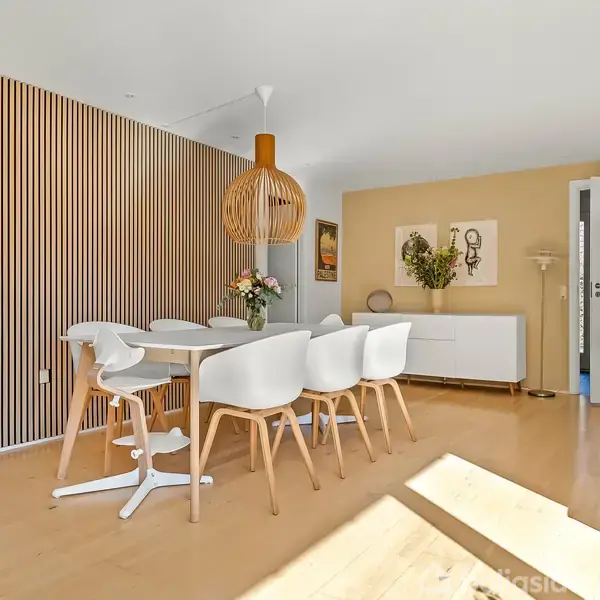 Spisebord med otte hvide stole står i et lyst rum med trægulv, moderne trævæg og kunst på sidevæggen. En stor pendellampe hænger over bordet.