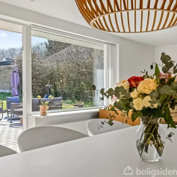 Blomster står i en vase på et hvidt bord. I baggrunden ses en lys stue med store vinduer, der vender ud mod en terrasse med møbler og have.