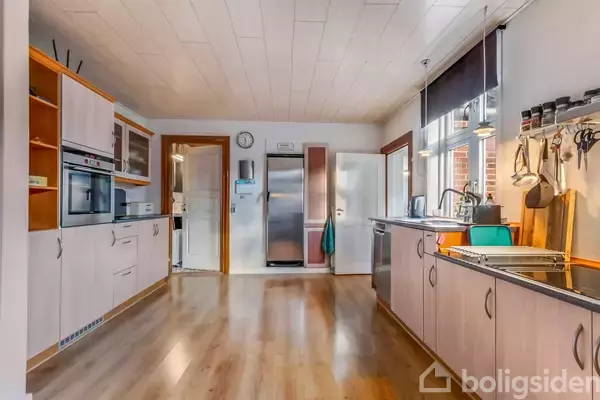 Køkken med trægulv, hvide skabe og sort bordplade. Indbygget ovn og mikrobølgeovn til venstre. Køleskab og ur på væggen. Vask ved vindue med redskaber hængende.