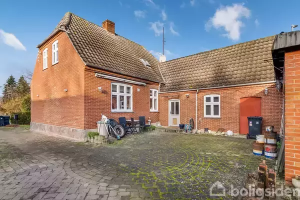 Et rødt murstenshus med tegltag står i en gårdsplads. Havemøbler og cykler ses ved siden af huset, omgivet af et brolagt areal. Himlen er blå med hvide skyer.