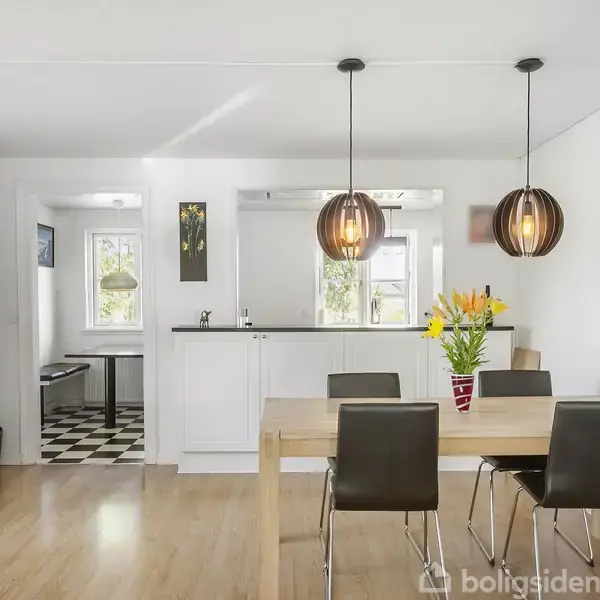 Spisebord med fire stole står i en moderne spisestue med lyst trægulv. Over bordet hænger to lamper, og en vase med gule blomster er placeret midt på bordet. I baggrunden er et køkken med sort-hvidternet gulv og hvide skabe.