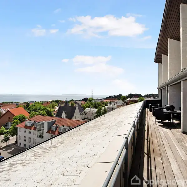Elevator står åben i lys lejlighed med sofa, stort tæppe og væg med vinduer, der viser balkon og udsigt over havet. Tekst: "Lavet med AI".