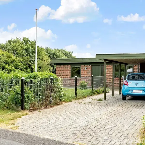 Blå bil holder parkeret i en carport foran et murstenshus. Huset er omgivet af grønne buske og træer. Et hegn og et vejskilt med tallet "18" ses i forgrunden.