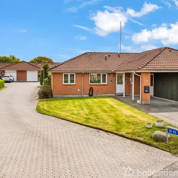 En rød murstensbungalow står med åben garage og anlagt græsplæne. En brolagt indkørsel fører til huset i et villakvarter. Skiltet ved vejen viser "19-23".