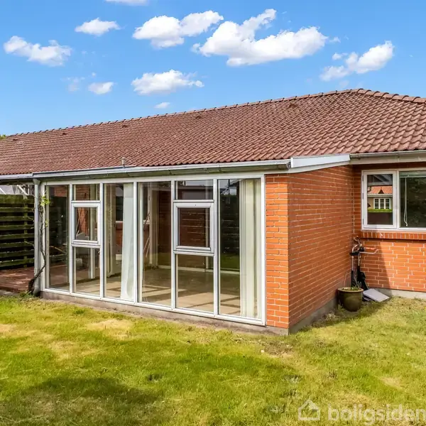 Et rødt murstenshus med et skråt tag har en udestue med store vinduespartier. Det hele ligger i en have med græsplæne og enkelte buske under en blå himmel med skyer.