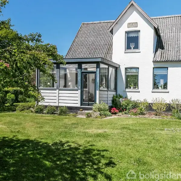 En hvid villa med en lille glaskonservatorium ved siden af. Villaen har en velplejet have og blå himmel i baggrunden.