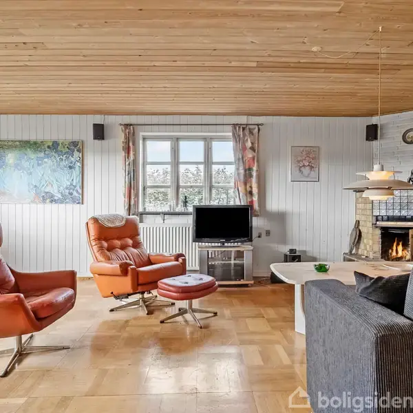 Brune lænestole og fodskammel står på et trægulv i en stue. En sofa og et tv er til venstre, mens en brændeovn brænder til højre, nær et vindue.