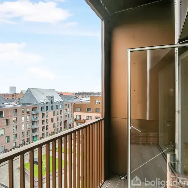 En balkon med glasskydedør åbner ud mod et bylandskab med moderne, grå og røde murstensbygninger under en skyet blå himmel.