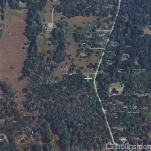 Luftfoto viser et skovområde med spredte huse og en snoet vej, der løber gennem landskabet. Omkringliggende vegetation er tæt med træer og skovklædte områder, hvilket skaber en landlig atmosfære.
