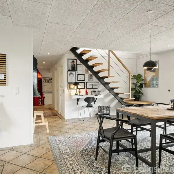 En spiseplads med et træbord og sorte stole står i fokus. Trappe til første sal i baggrunden, omgivet af kunst på væggen og naturligt lys fra store vinduer.