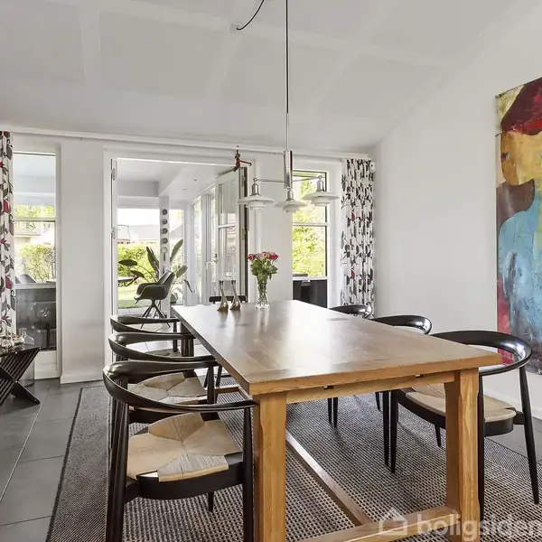 Et spisebord i træ med sorte stole omkring står i et moderne rum. En stor, farverig kunstmaleri hænger på væggen. Store vinduer med blomstrede gardiner giver lysindfald.
