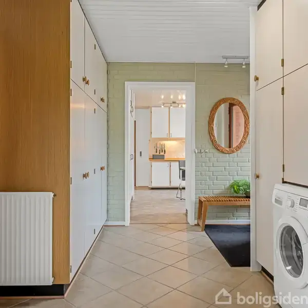 Vaskemaskine står i en gang med hvide skabe og en radiator på væggen. Der er et rundt spejl og en bænk. Gangen fører til et rum med køkkenelementer.
