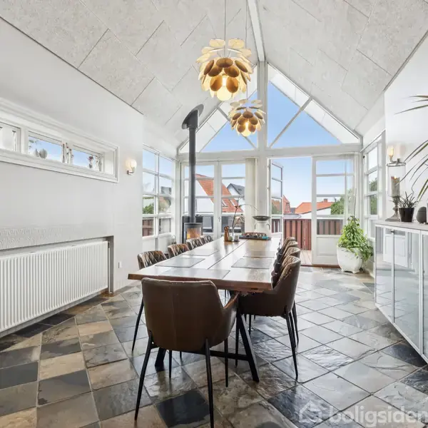 Et spisebord med stole står i en lys stue med højt til loftet, store vinduer og skifergulv. Plantepynt og moderne lamper skaber en hyggelig atmosfære.