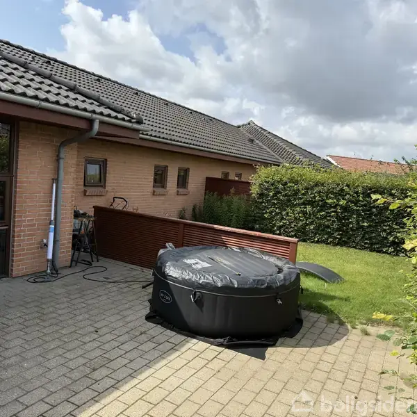 Et oppusteligt spabad står på en flisebelagt terrasse ved siden af et murstenshus med tagsten. I baggrunden er der en velplejet have med grønne buske.