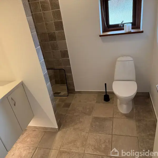 Toilet står i et badeværelse, flankeret af en toiletbørste og vindue ovenfor. Ved siden af er en vask med underskabe og bruseniche med grå fliser.