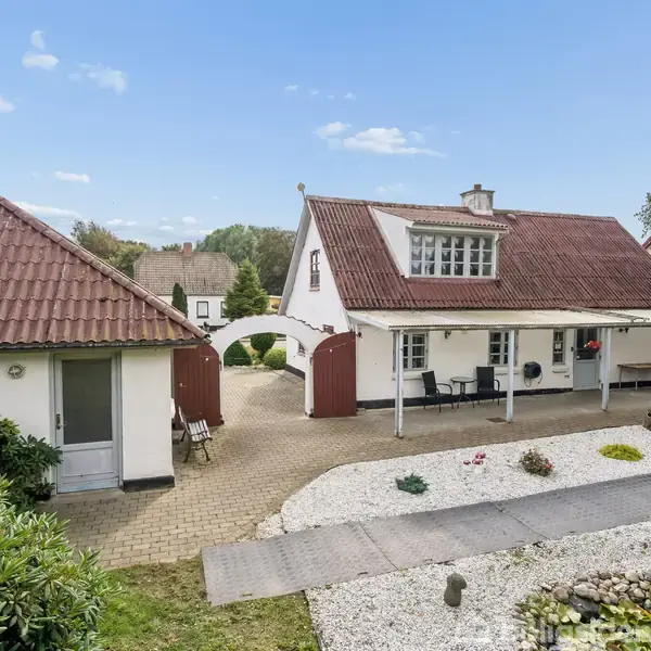 Et hvidt hus med rødt tegltag og en veranda står i en velplejet gårdsplads med stenbelægning. Der er også en mindre bygning til venstre, omgivet af frodig vegetation.
