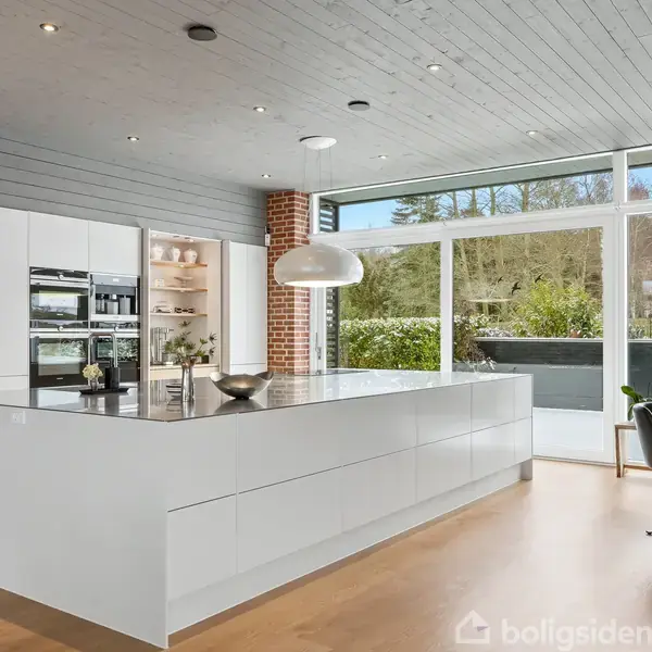 Et moderne køkken med en stor hvid køkkenø, indbyggede ovne i hvide skabe, og en stor lampe hængende over øen. Store vinduer giver udsigt til en terrasse og have.
