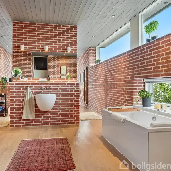 Håndvask hænger på en rød murstensvæg i et rummeligt badeværelse med træloft og store vinduer. Et badekar står ved et vindue med udsigt til grønne planter.