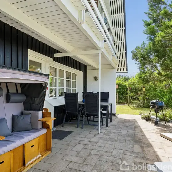 Udendørs terrasse: En strandkurv og havemøbler står på en flisebelagt terrasse ved et sortmalet hus. En grill er placeret ved siden af, omgivet af grønne buske og træer.