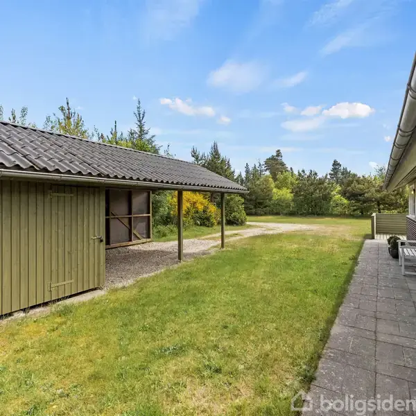 Garagebygning med grønt tag står til venstre, stille på en græsklædt grund. Til højre ses en bygning med terrasse, omgivet af træer og buske under blå himmel.