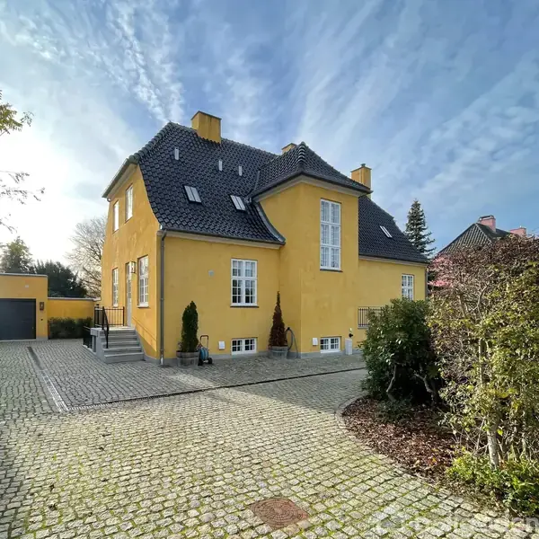 Gult hus med sort tegltag og hvide vinduer, placeret midt i en brolagt indkørsel omgivet af hække og mindre træer under en blå og let skyet himmel.