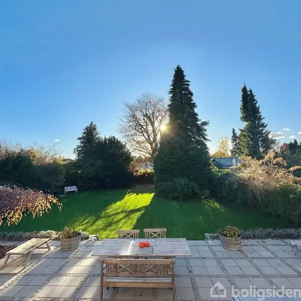 En udendørs terrasse med et bord og bænke står på fliser, omgivet af græsplæne og høje træer. Solen skinner mellem træerne i en have på en klar dag.