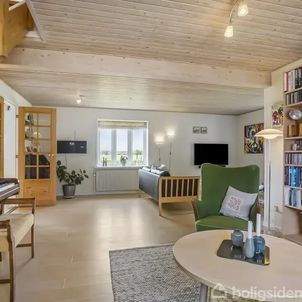 Et gult hus med rødtag omgivet af en hække- og træbevoksning, står ved en grusvej i et landligt område med åbne marker i baggrunden.