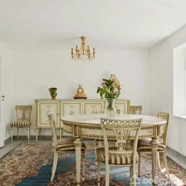 Et elegant spisebord med stole står på et orientalsk tæppe. Rummet har en hvid kommode, blomsterdekorationer og en lyskrone. Store vinduer giver naturligt lys.