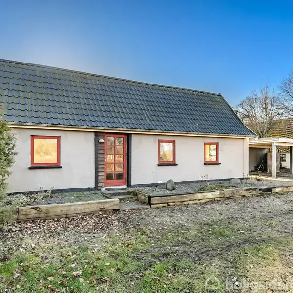 Et lille, hvidt hus med røde vinduesrammer og sort tegltag står i en have. Foran huset er en lille haveplæne med bede og et grusareal.