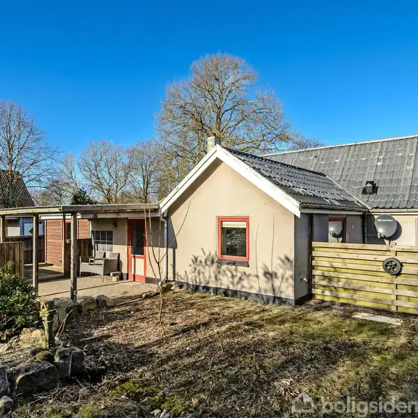 Hus står stille i have med græsplæne og buske; omgivet af træer og blå himmel; har røde vinduesrammer, gråt tag og terrasseområde til højre.