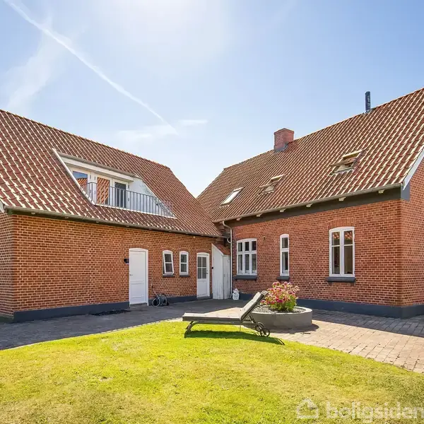 Et rødt murstenshus med et skråt tegltag står i et solrigt gårdmiljø. En liggestol og blomsterkasser ses på græsplænen foran huset, omgivet af en flisebelagt terrasse.