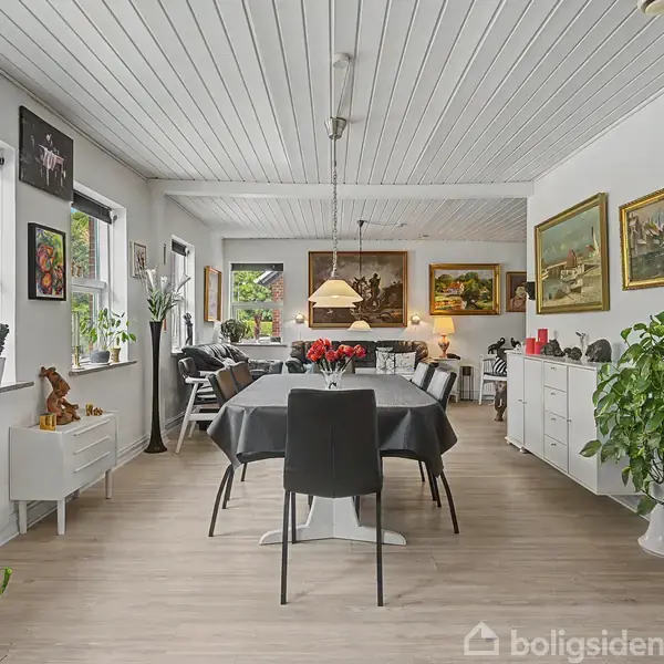 Spisestue med langt bord omgivet af sort-hvide stole. Bordet er dækket med en sort dug og vase med røde blomster. Væggene er dekoreret med malerier. Planter står langs vinduerne.