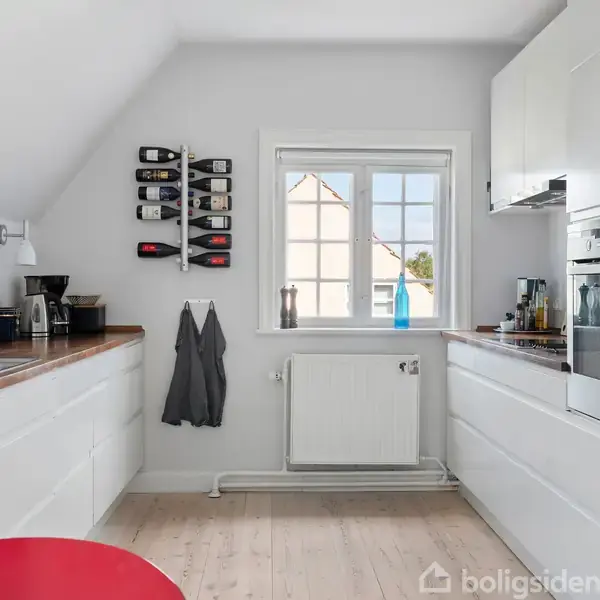 Køkken med hvide skabe og træbordplade indeholder en ovn, kaffemaskine og vase. Et vindue lyser rummet. En rød stol står centralt. Vinhylde og viskestykker hænger på væggen.