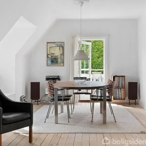 Et spisebord med fire stole står på et tæppe i et lyst rum. En dør åben til en terrasse med grøn udsigt. Værelset har moderne kunst på væggene.