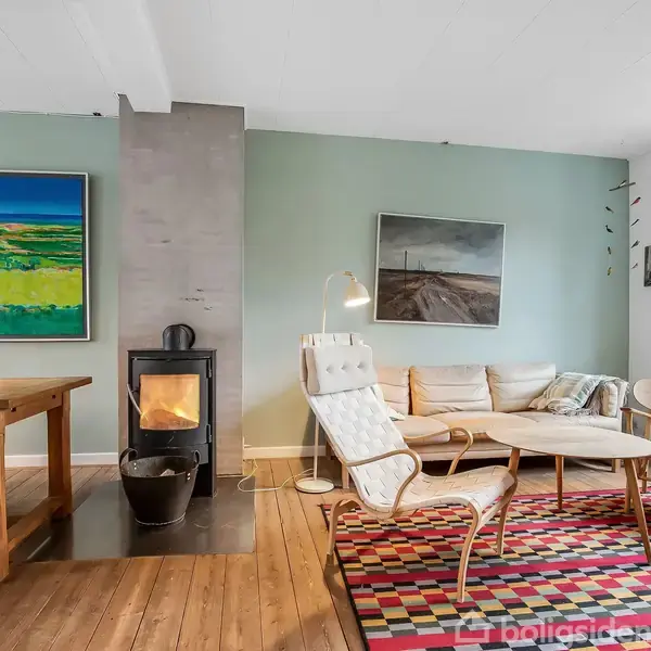 Stue med brændeovn i brug, omgivet af en lænestol, sofabord, sofa og spisebord. Farverigt tæppe på trægulv, malerier på væggene; lysegrønne vægge og vindue til højre.