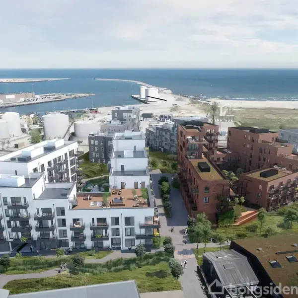 Lejlighedskomplekser står stille ved kysten, hvor de omkranses af grønne områder og havet i baggrunden. Industribygninger kan ses til venstre tæt ved havneområdet.