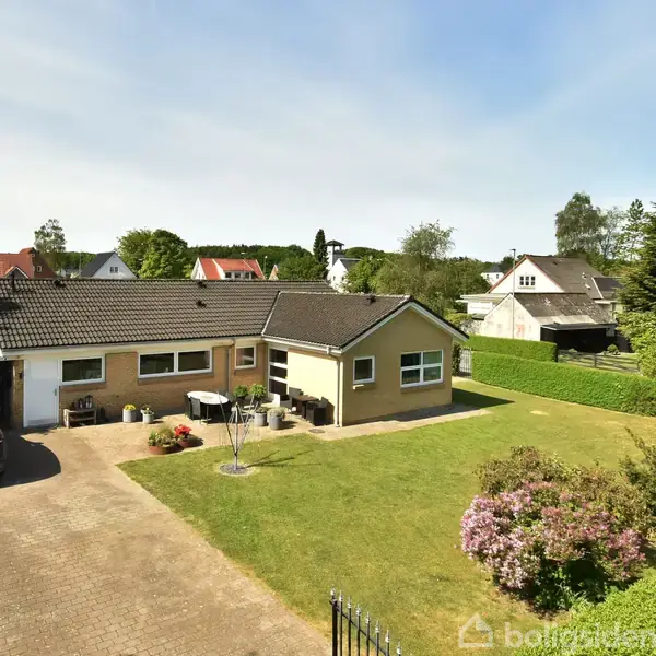 Et gult parcelhus med mørkt tag står omgivet af en velplejet have med blomsterbede. En bil er parkeret i en indkørsel ved siden af huset.