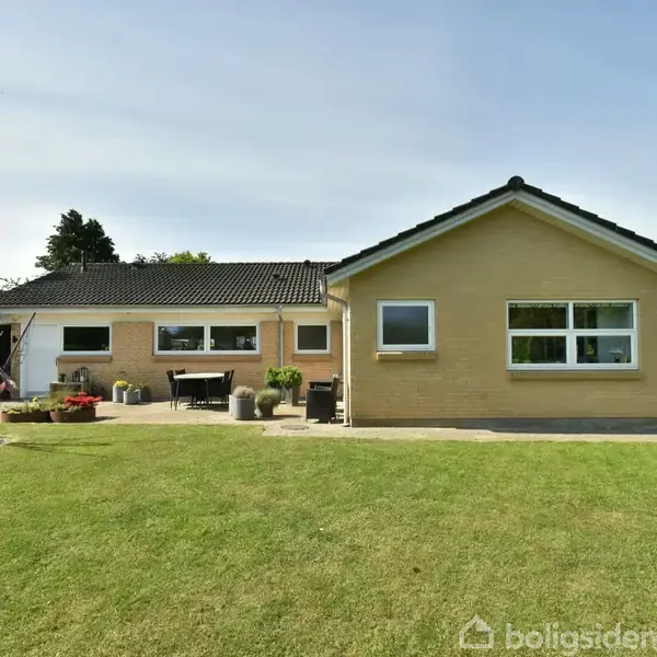 Et enkelt etplanshus med gul murstensfacade og sorte tagsten står i en velplejet have. En terrasse med borde og stole er synlig, omkranset af græsplæne og træer.