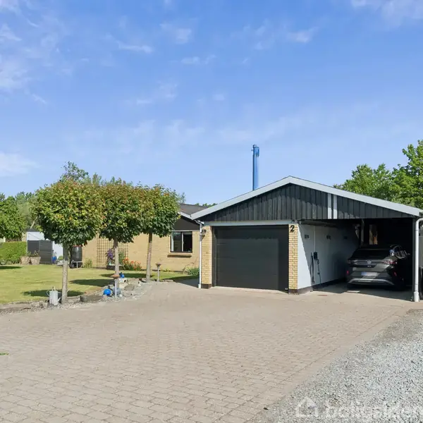 Garagebygning står ved siden af et enfamiliehus, med en bil parkeret i en åben carport. Flisebelagt indkørsel omgivet af græsplæner og træer, under en klar blå himmel.