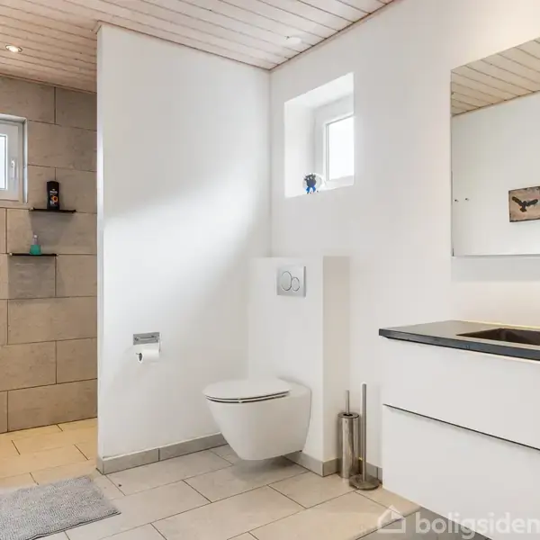 Et lyst badeværelse med hængende toilet og moderne vask. Blåt håndklæde hænger ved siden af brusekabinen. Rummet har lyse fliser og en spejl over vasken. Tre små billeder hænger på væggen.