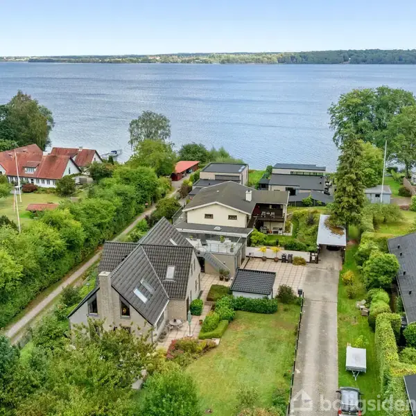 En række huse ligger langs en vej ved vandet, omgivet af frodige haver og træer. I baggrunden ses en stor vandstrækning med en skov på den anden side.