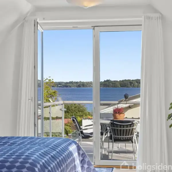 Soveværelse med blåternet sengetæppe fører ud til en balkon gennem glasdøre. På balkonen står et bord og stole med udsigt over en sø og skovklædte bakker i baggrunden.