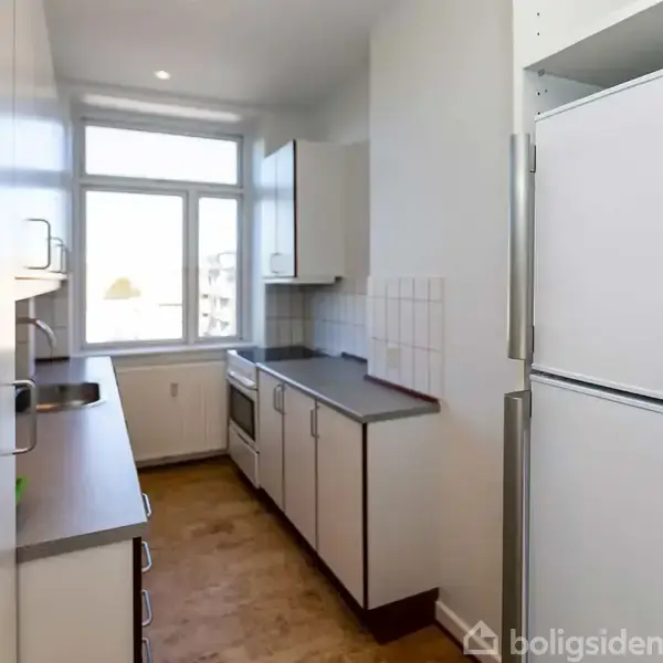 Et smalt køkken med hvide skabe og en ovn ved siden af et vindue. Et køleskab står i forgrunden, mens en lysstrøm kommer ind fra vinduet.