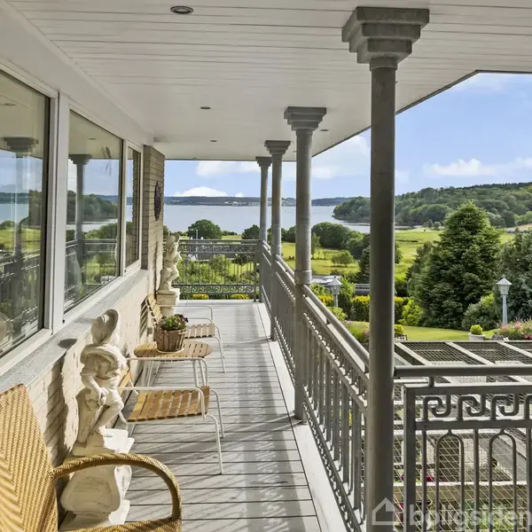 En overdækket altan med kurvestole og dekorative figurer står ud mod en have med træer og udsigt til en sø i baggrunden.