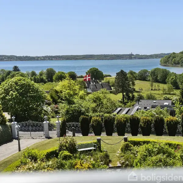 En haver med en bænk står i forgrunden, mens en dansk flag vajer over en ejendom omringet af frodig skov. I baggrunden strækker en rolig sø sig under en klar blå himmel.