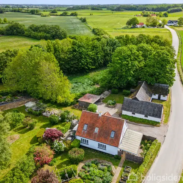 Hus med rødt tag og hvide vægge, omgivet af have og grønt, ligger langs snoet landevej i landligt område med marker og træer i baggrunden.