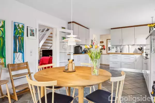 Et rundt træbord med fire stole er centralt placeret i et lyst køkken. En vase med tulipaner står på bordet, omgivet af moderne hvide skabe.