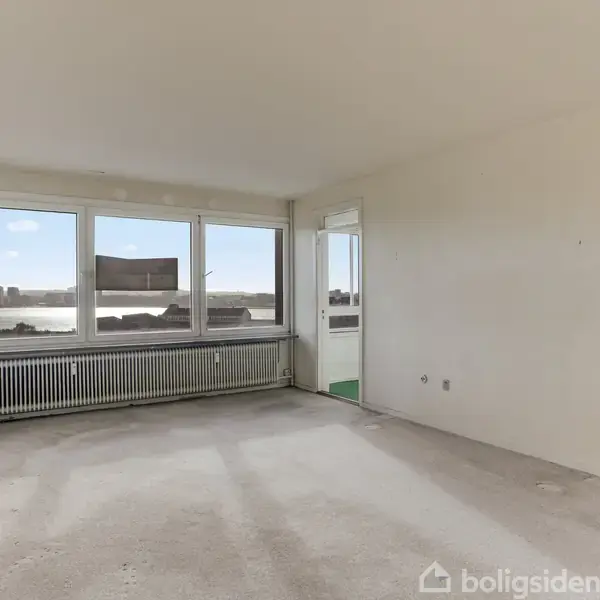 Et lyst, tomt rum med store vinduer og udsigt over en by og en flod. Der er en radiator under vinduerne og en dør til højre, der fører til et andet rum.
